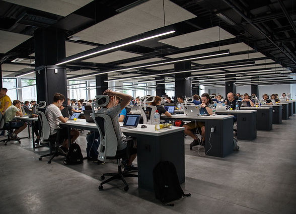 An open-plan workspace in daylight: long desk runs, task chairs, and a concrete floor beneath an exposed ceiling.